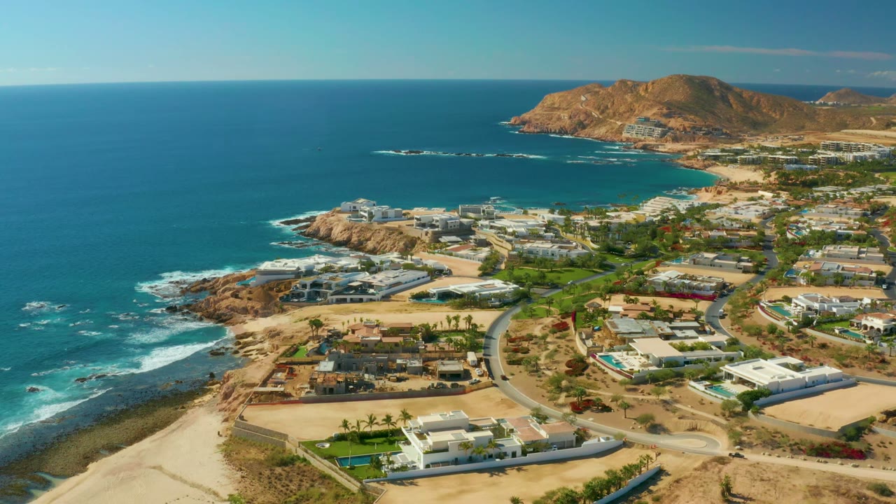 Wide drone view of waves breaking beside coastal homes and roads.