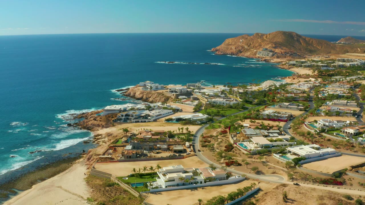 Aerial frame over Chileno Beach, Los Cabos, with the blue bay extending across the upper frame, surf wrapping a rocky point, residential streets below, distant hills, and no visible people.