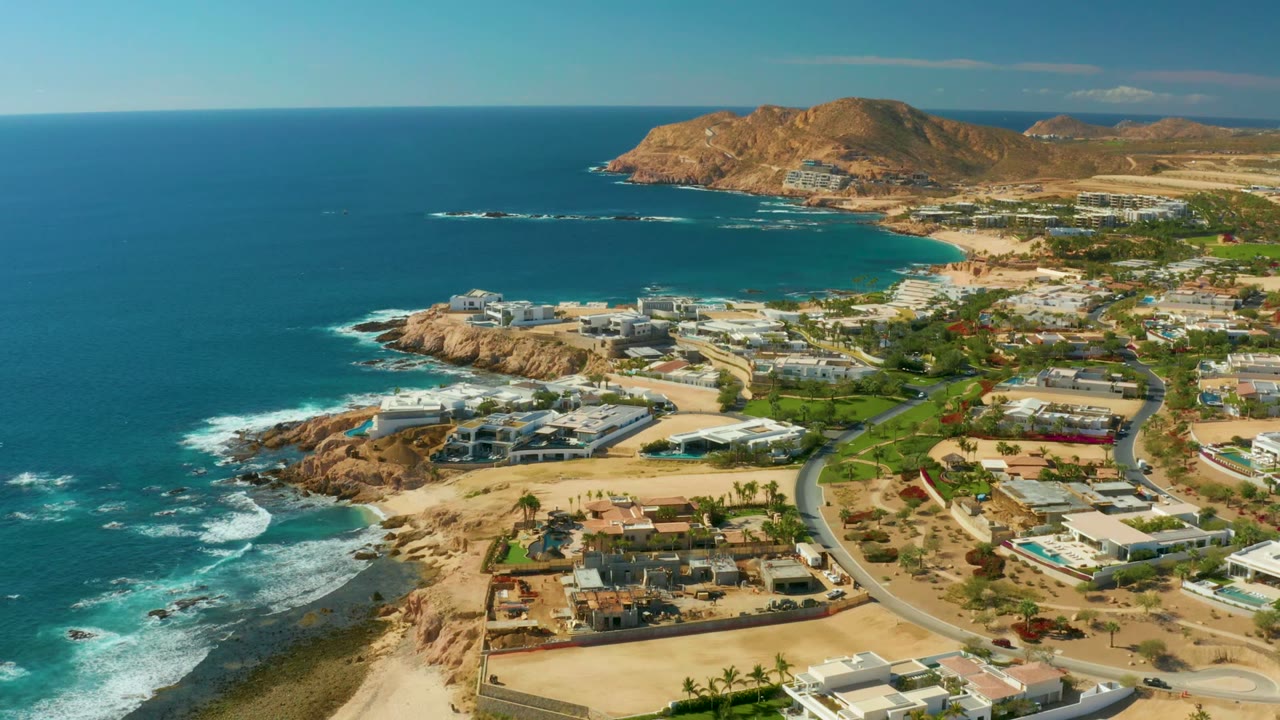 Aerial frame over Chileno Beach in Los Cabos showing blue ocean on the left, rocky shoreline, a curving coastal road, modern buildings, dry hills, and no visible people.