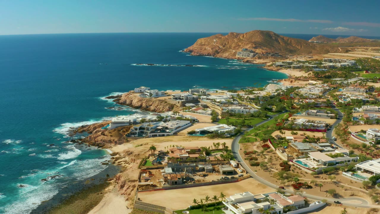 Wide drone view of coastal buildings, road traffic, surf, and arid hills.