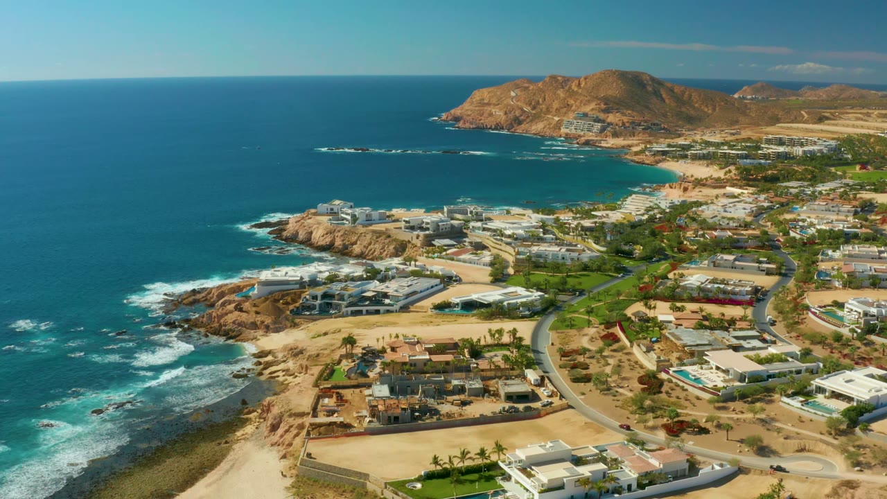 Aerial panorama at Chileno Beach showing turquoise and deep blue water, low oceanfront villas, a winding road, sandy lots, rocky points, distant hills, and no visible people.