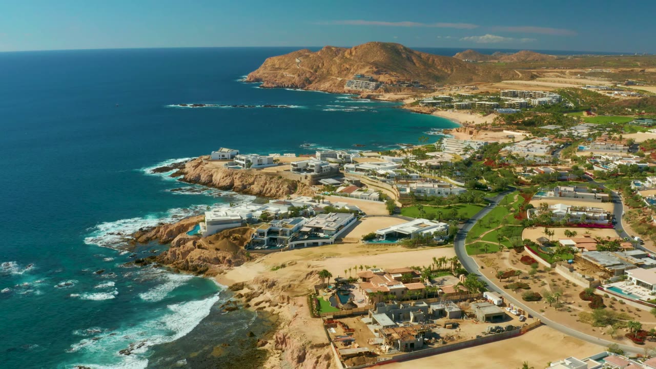 Opening aerial frame over Chileno Beach Los Cabos with blue ocean on the left, white surf on rocks, coastal homes, roads, sandy ground, distant hills, and no visible people.