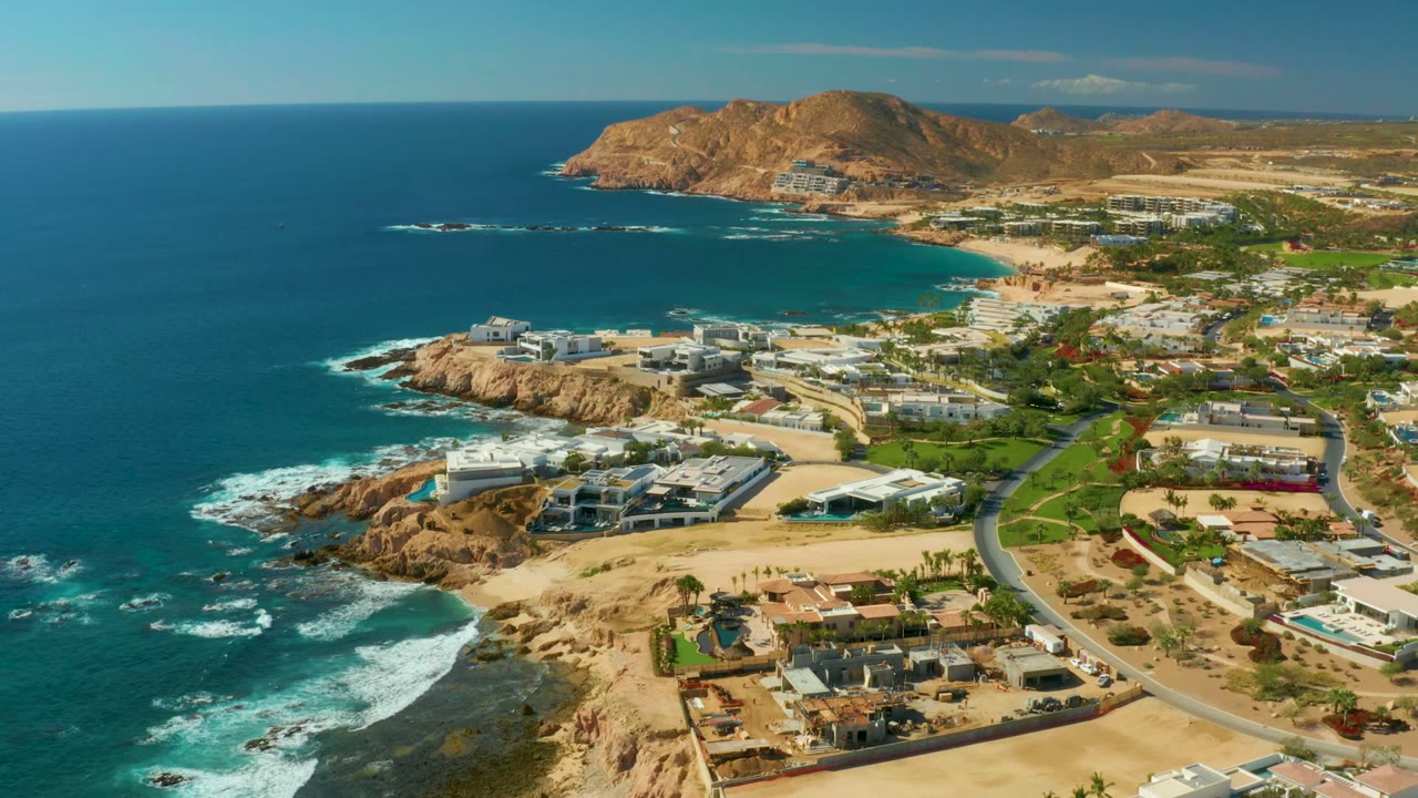 Wide drone view of rocky shoreline, ocean waves, residences, roads, and hills.