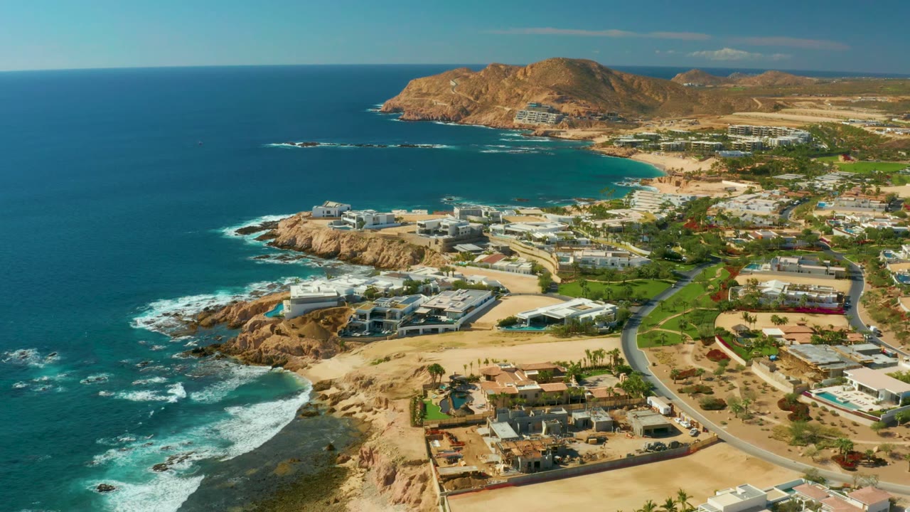Aerial frame from Chileno Beach Los Cabos with turquoise water, surf lines along the rocky points, a small sandy beach, modern homes and roads inland, brown hills in the distance, and no visible people.