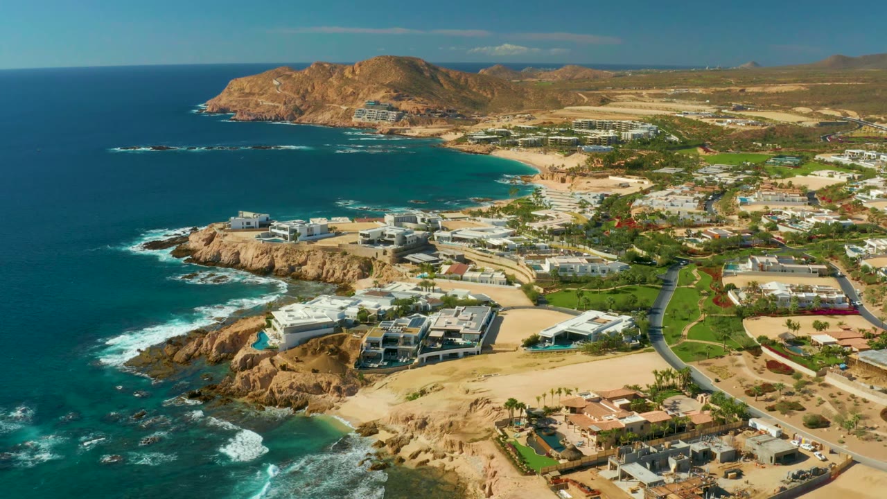 Opening aerial frame over Chileno Beach Los Cabos with blue ocean on the left, white surf hitting rocks, cliffside buildings, a curving road, brown hills in the distance, and no visible people.