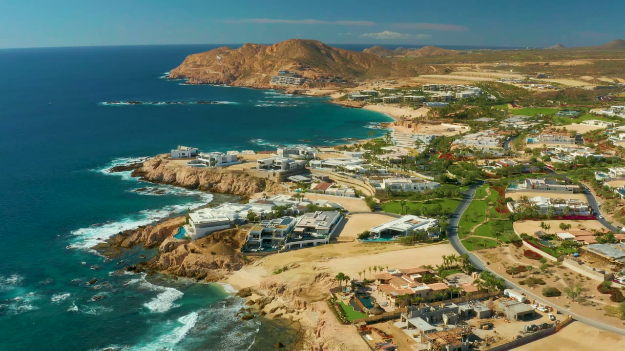 At 0:02, the Chileno Beach Los Cabos coastline remains in a wide aerial composition with whitecaps near the lower rocks, sandy lots and villas across the center, road curves on the right, and no visible people.