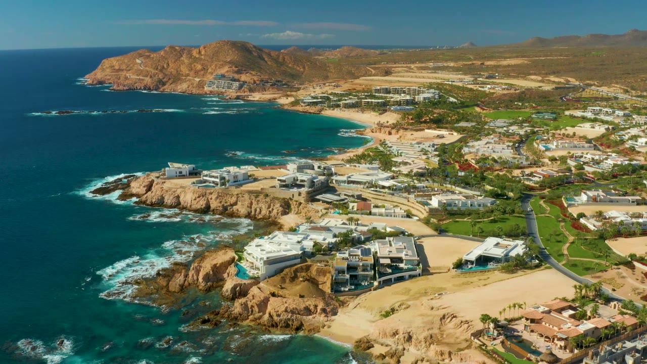 Opening aerial frame over Chileno Beach Los Cabos with blue ocean on the left, surf around tan rocks, beachfront buildings and inland hills visible, with no visible people.