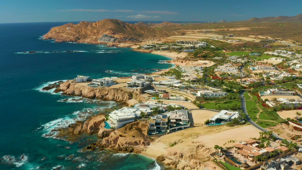 Ocean waves break against rocks beside beachfront buildings and hills.
