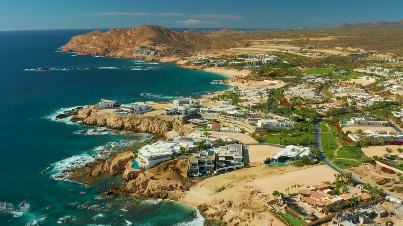Aerial frame at 0:02 over Chileno Beach Los Cabos with turquoise water curving into the beach, surf wrapping the rocks, clustered white buildings, inland greenery, and no visible people.