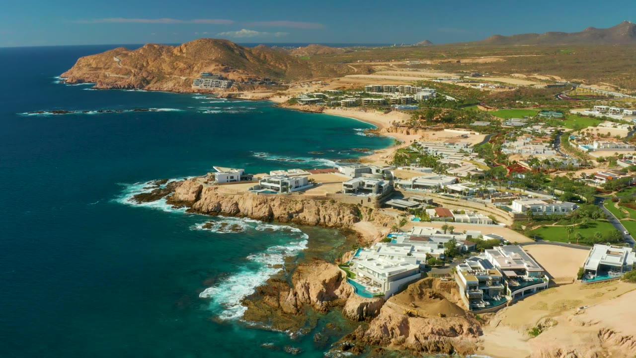 Opening aerial frame over Chileno Beach in Los Cabos, showing dark blue ocean, tan cliffs, white shoreline buildings, inland hills, and no visible people.