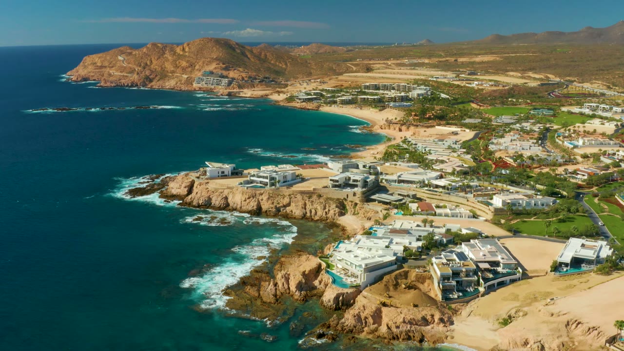 High coastal view with surf around rocks and white buildings near the shore.