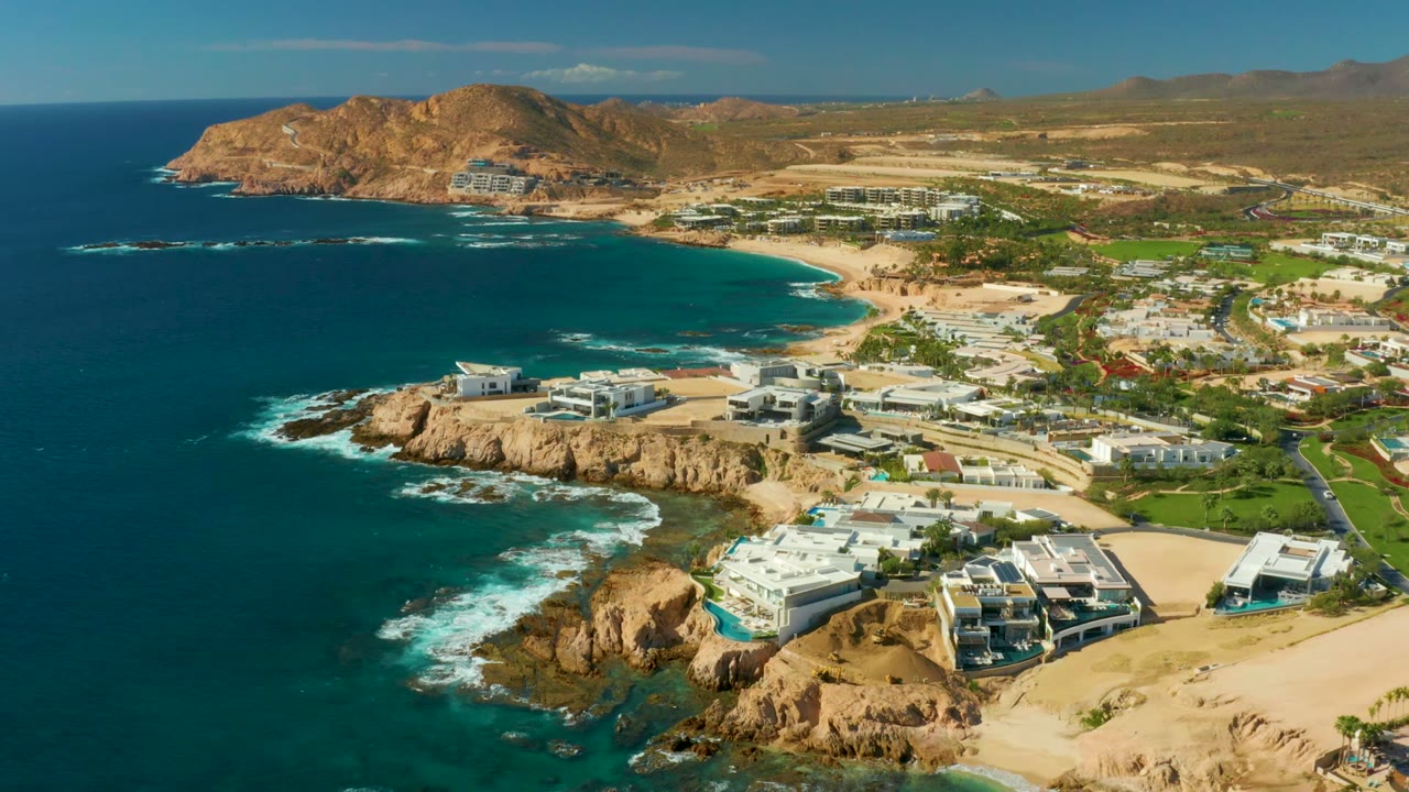 Wide drone-like frame over Chileno Beach Los Cabos, with the bay on the left, developed shoreline on the right, dry hills in the distance, and no visible people.