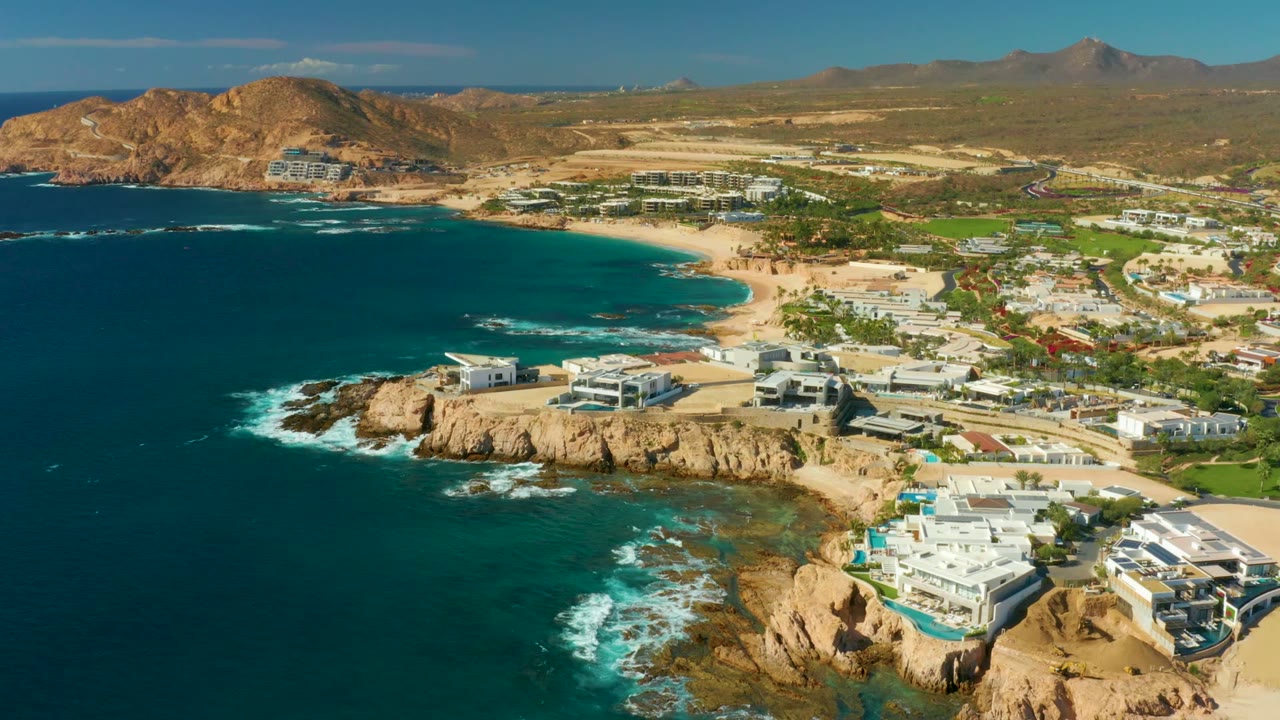 Aerial frame over Chileno Beach Los Cabos showing deep blue ocean at left, tan rocky cliffs, white cliffside buildings, a blue pool, sandy beach, dry hills, and no visible people.