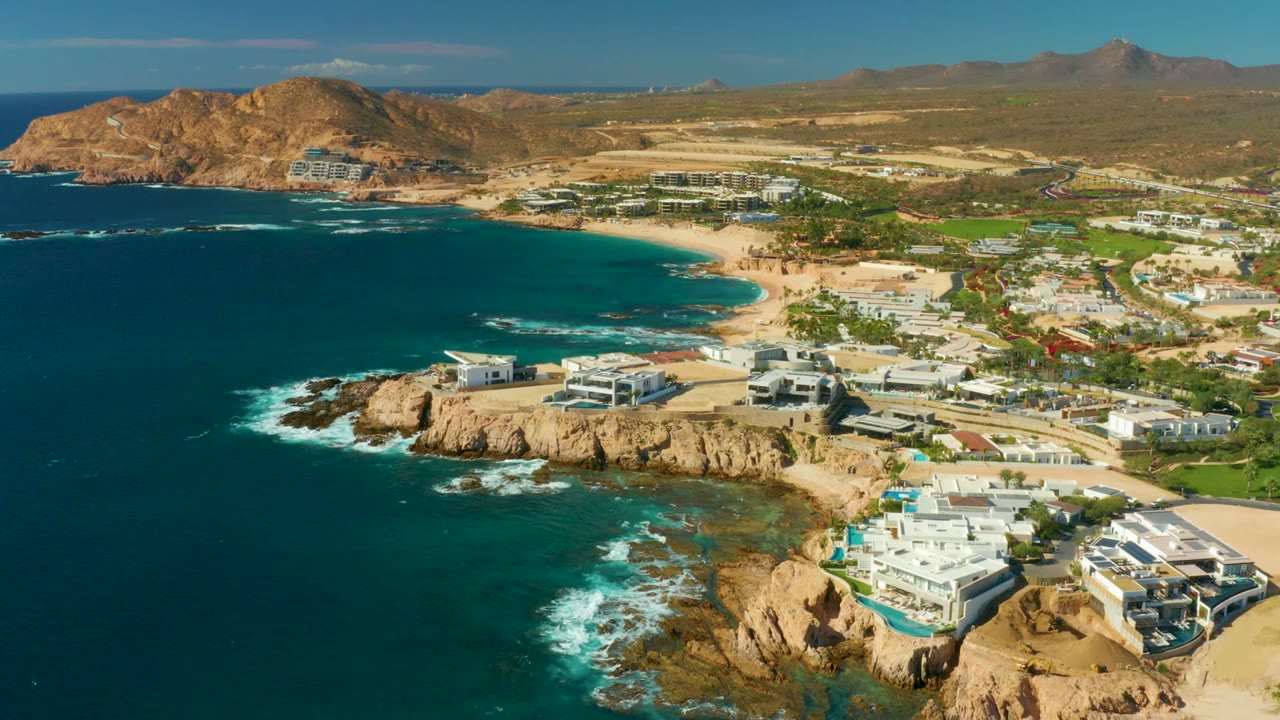Wide aerial coastline with ocean, rocky point, beach, and modern white buildings.