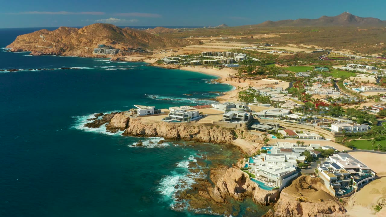 Aerial frame over Chileno Beach Los Cabos with turquoise water curving into a sandy beach, white buildings on tan cliffs in the foreground, arid hills behind, and no visible people.