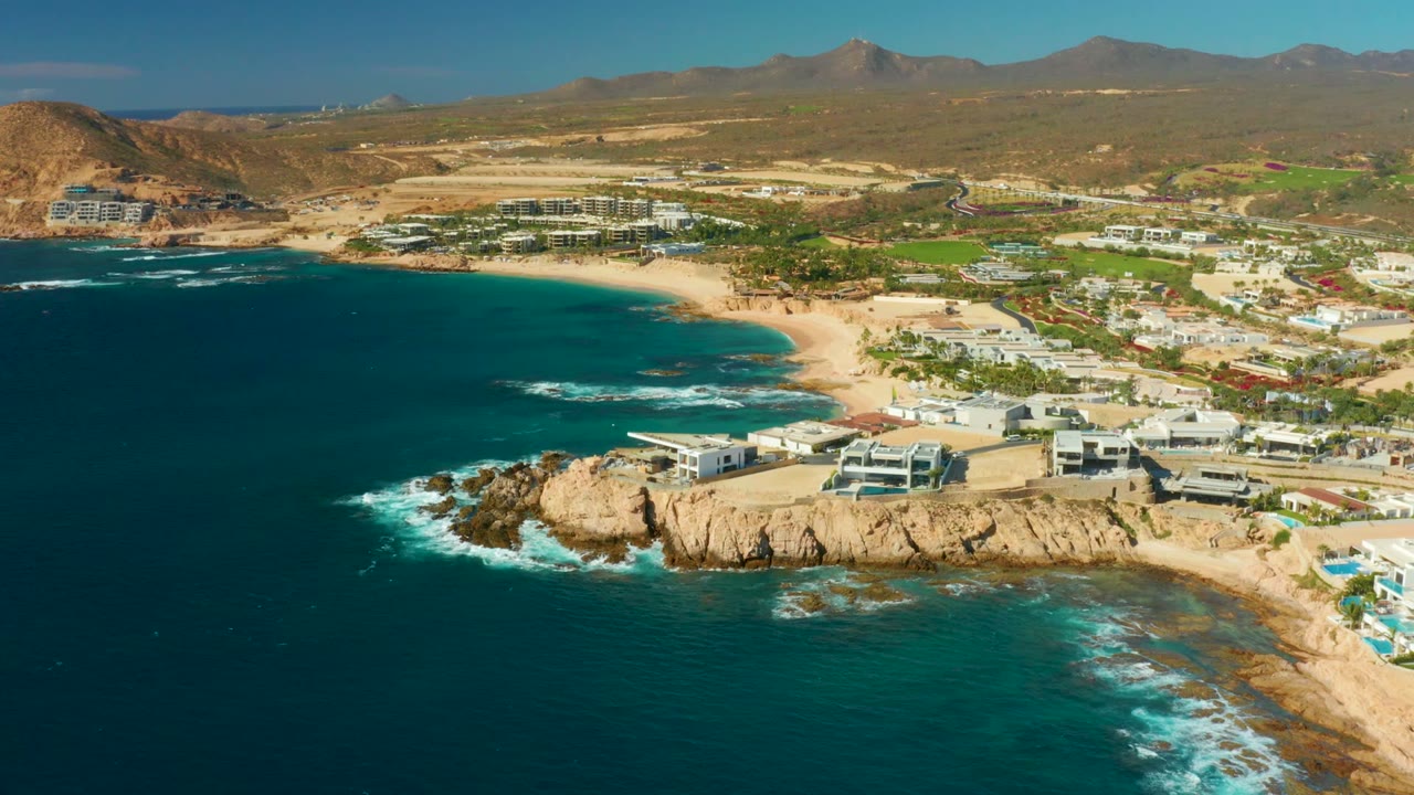 Opening aerial frame over Chileno Beach Los Cabos with dark blue ocean in front, tan rocky cliffs, a sandy beach, white resort buildings, dry hills, distant mountains, and no visible people.