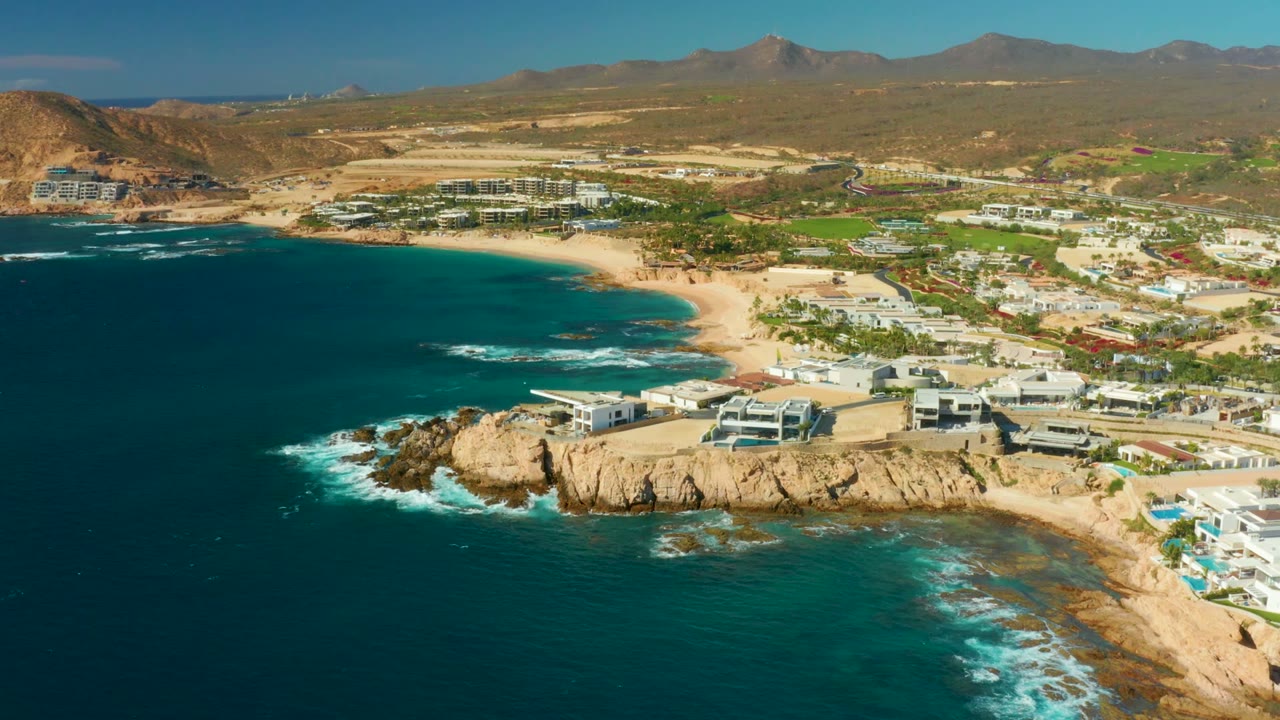 Drone view of a rocky point, turquoise shallows, sandy beach, white buildings, and inland mountains.