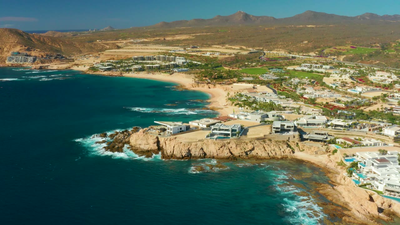 Wide aerial frame over Chileno Beach Los Cabos with ocean filling the lower half, waves breaking beside tan cliffs, beach and resort buildings across the middle distance, mountains on the horizon, and no visible people.