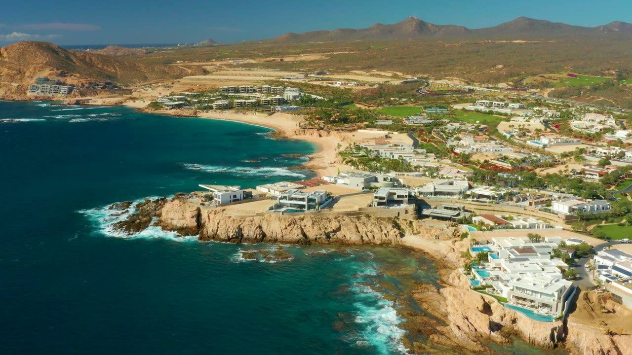 Opening aerial frame over Chileno Beach in Los Cabos, with turquoise water on the left, surf striking tan rocks, beachfront buildings on the right, arid hills beyond, and no visible people.