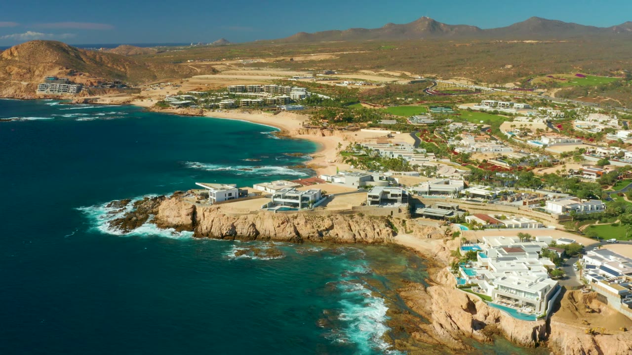 Wide drone view of ocean waves breaking below coastal buildings and beach