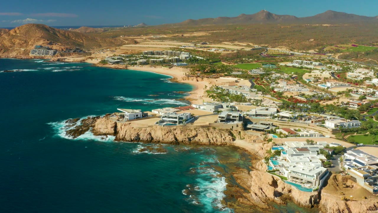 Chileno Beach aerial view with blue-green shallows around the rocks, a sandy cove curving into the center, modern coastal buildings to the right, dry hills behind town, and no visible people.