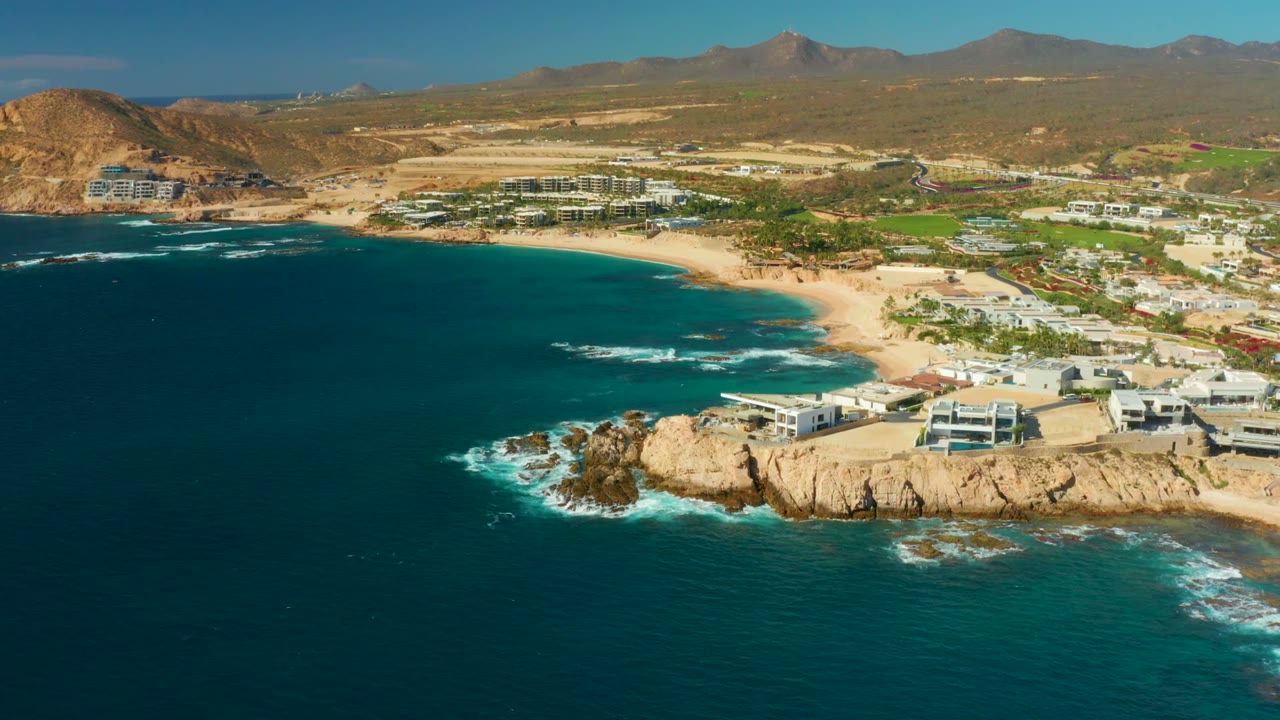 Aerial view of Chileno Beach in Los Cabos with dark blue water in the foreground, a rocky point with low buildings, sandy shoreline, distant dry mountains, and no visible people.