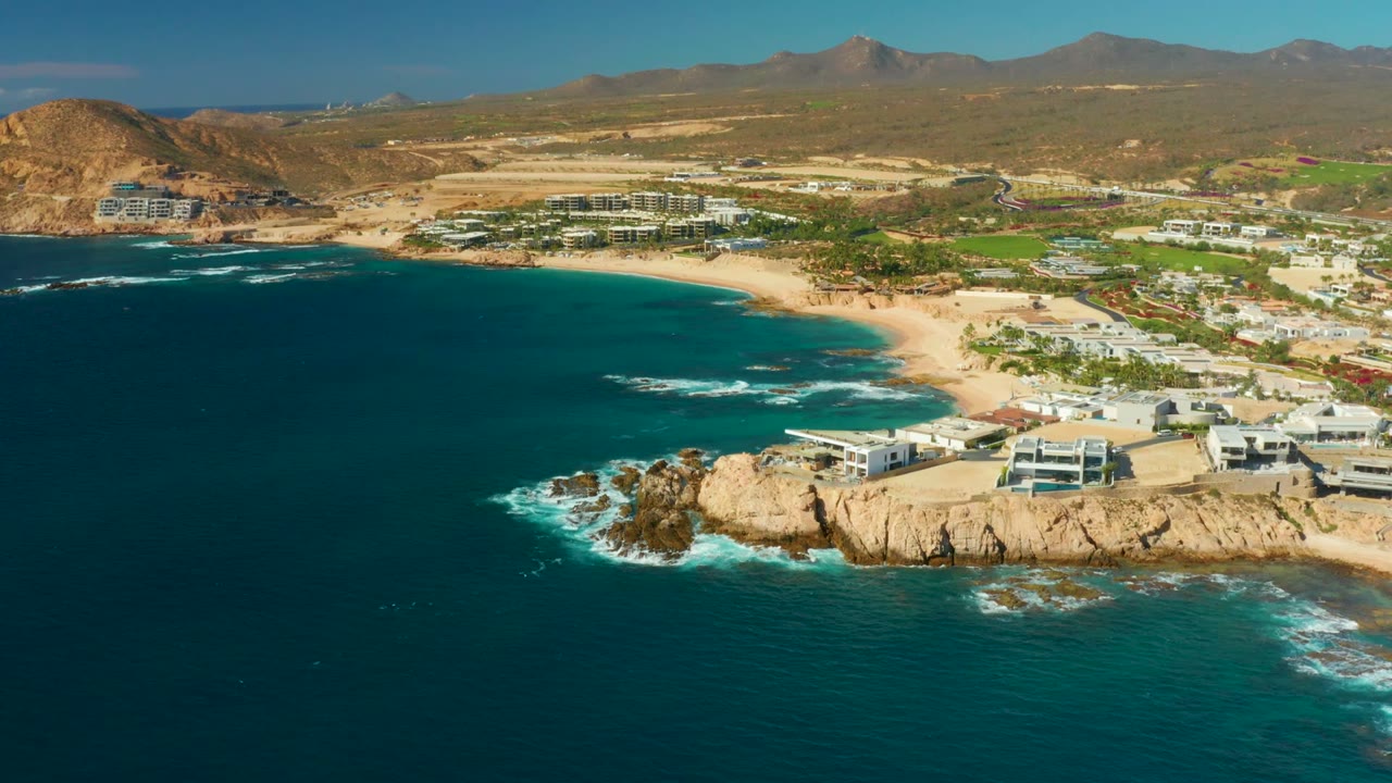 Aerial coastal view with ocean waves around rocks and beachfront buildings near sand.