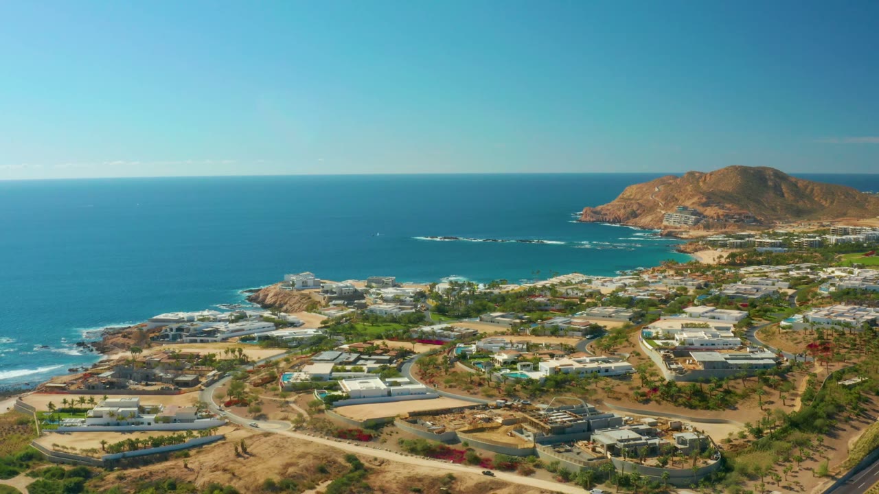 Opening aerial frame over Chileno Beach Los Cabos shows blue ocean, a rocky point, low coastal buildings, curving roads, dry terrain, and no visible people.