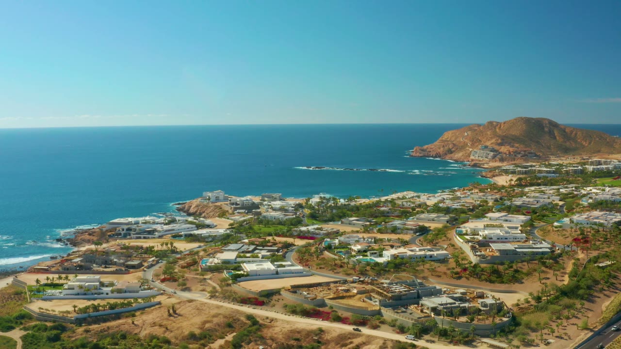 At 0:02, the frame shows Chileno Beach Los Cabos from above with open sea on the left, a rocky headland to the right, built coastal lots, roads, and no visible people.