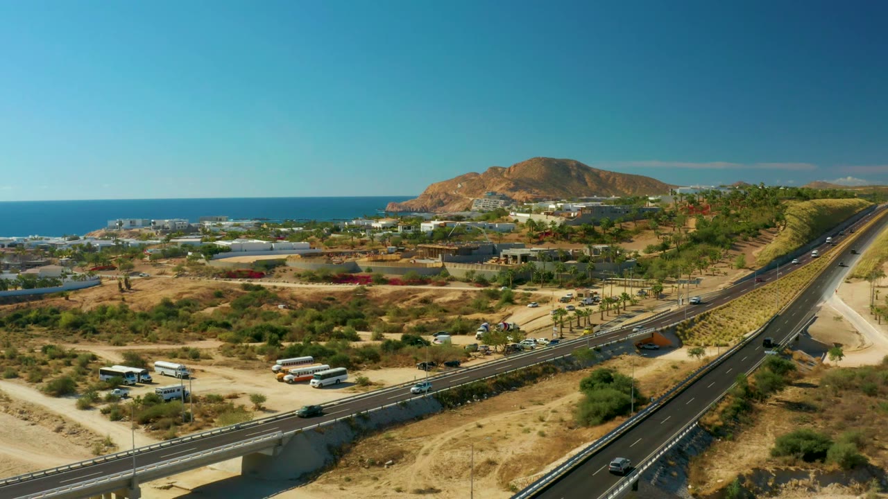 At 0:01 in Chileno Beach Los Cabos, vehicles are spaced along the highway lanes and bridge, with a white truck area, palms, low structures, ocean, ridge, and no visible people.