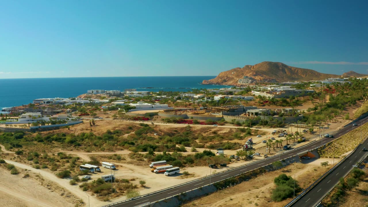 Aerial frame at Chileno Beach Los Cabos with cars and buses near the lower road area, the highway crossing the right side, ocean beyond buildings, and no visible people.