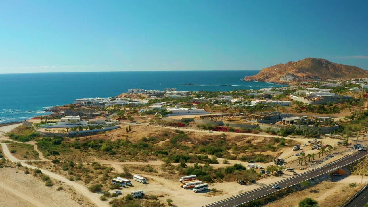 Opening aerial frame over Chileno Beach Los Cabos with dry foreground tracks, an asphalt road, coastal buildings, blue ocean, distant hills, and no visible people.