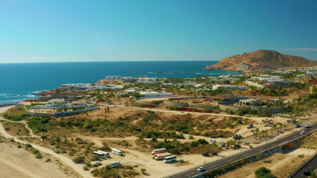 Wide drone view of coastal buildings beside ocean and hillside terrain.