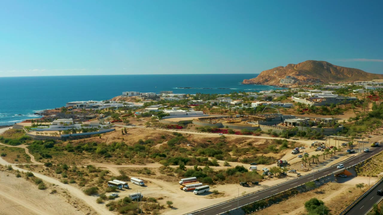 At 0:02, the drone perspective shows Chileno Beach Los Cabos with cars along the lower road, pale dirt tracks, clustered buildings, blue water, hills, and no visible people.