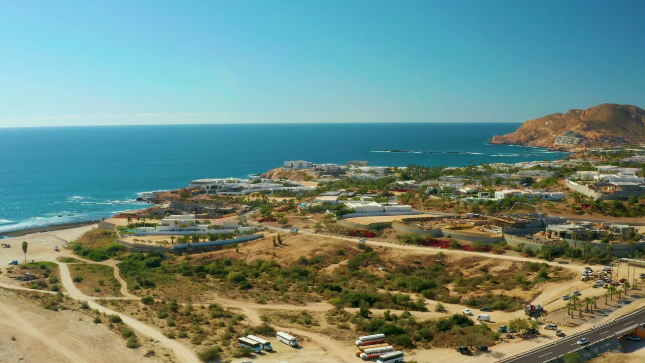 Opening aerial frame over Chileno Beach Los Cabos with sandy tracks in front, a coastal road at lower right, ocean and surf to the left, hillside buildings beyond, and no visible people.