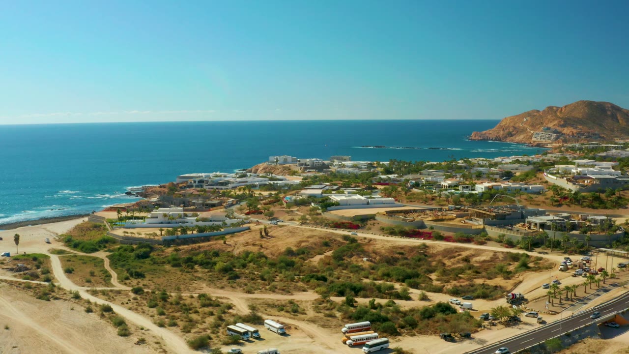 Aerial coastal view with blue ocean, beach access tracks, road traffic, buildings, and hill.