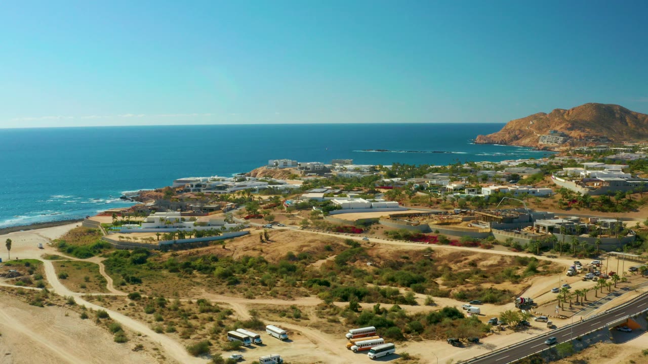 At 0:02, the frame shows Chileno Beach Los Cabos from above with the ocean stretching behind beachside roads, scattered low buildings, a rocky hill to the right, and no visible people.