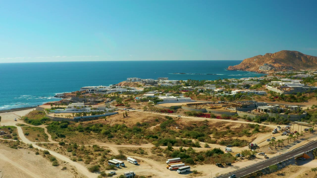 Wide aerial of coastal terrain, road traffic, beach, ocean, buildings, palms, and hill.