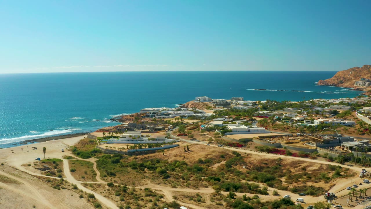Wide aerial view of a coastal road, sandy beach, buildings, dry hills, and blue ocean.