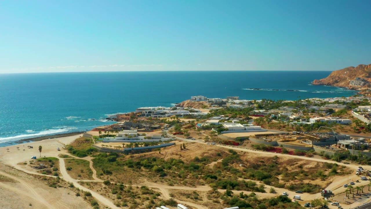 Chileno Beach Los Cabos aerial frame with vehicles along the coastal road, pale sand tracks in the foreground, low buildings, blue water, and no visible people.