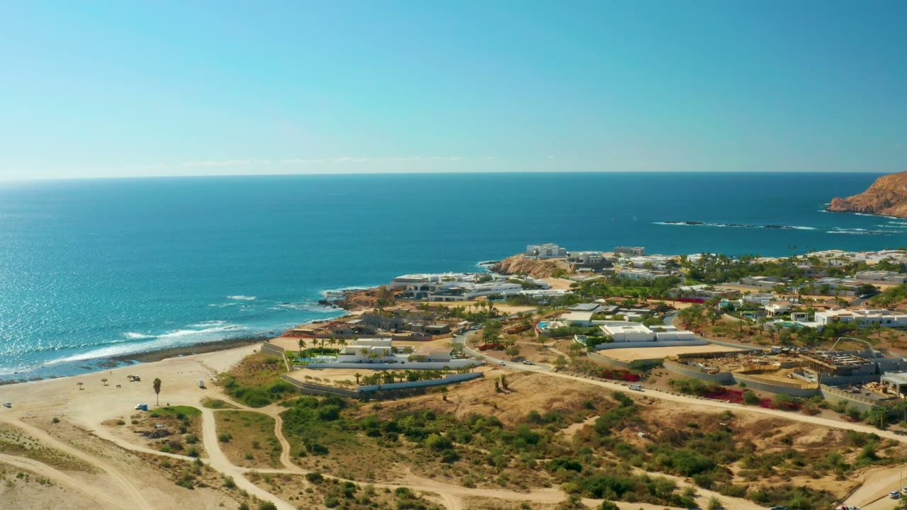 Aerial view over Chileno Beach in Los Cabos with blue ocean at left, sandy shore below, dirt paths crossing dry hillside, coastal buildings in the midground, and tiny people visible.