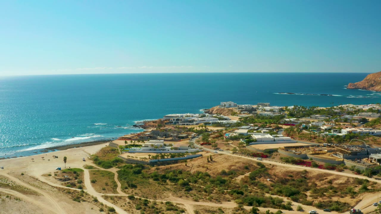 Chileno Beach aerial view with bright blue water on the left, beach and surf line below, winding dirt roads across the hillside, scattered modern buildings, and small people visible along the coastal area.
