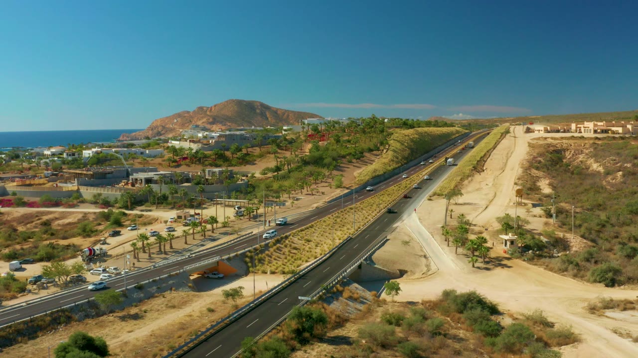 Aerial view of a divided coastal road with cars, dry terrain, palms, ocean, and hill.