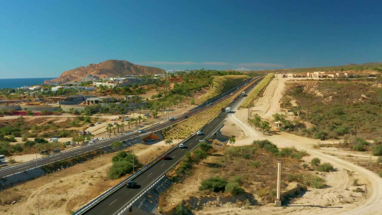 Aerial view of a coastal highway with cars, dry hills, ocean, and blue sky.