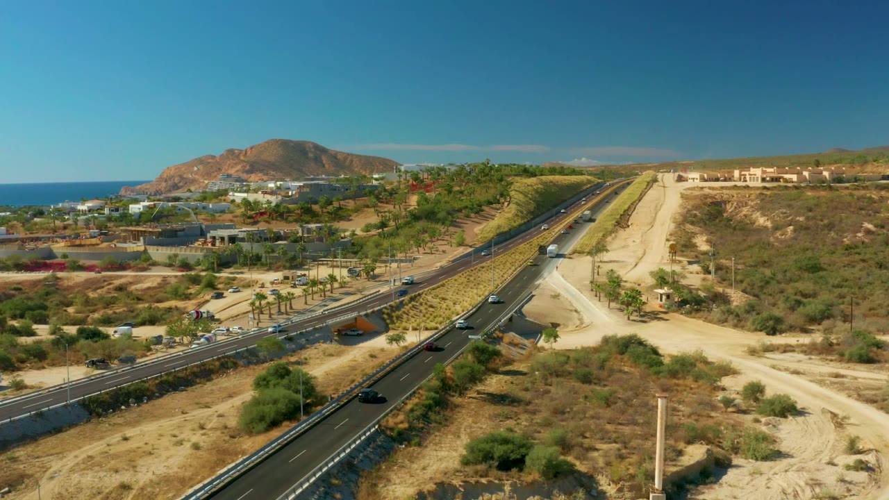 Aerial frame at one second showing Chileno Beach Los Cabos highway traffic, a dirt access road, sparse vegetation, ocean horizon, hills, and no visible people.