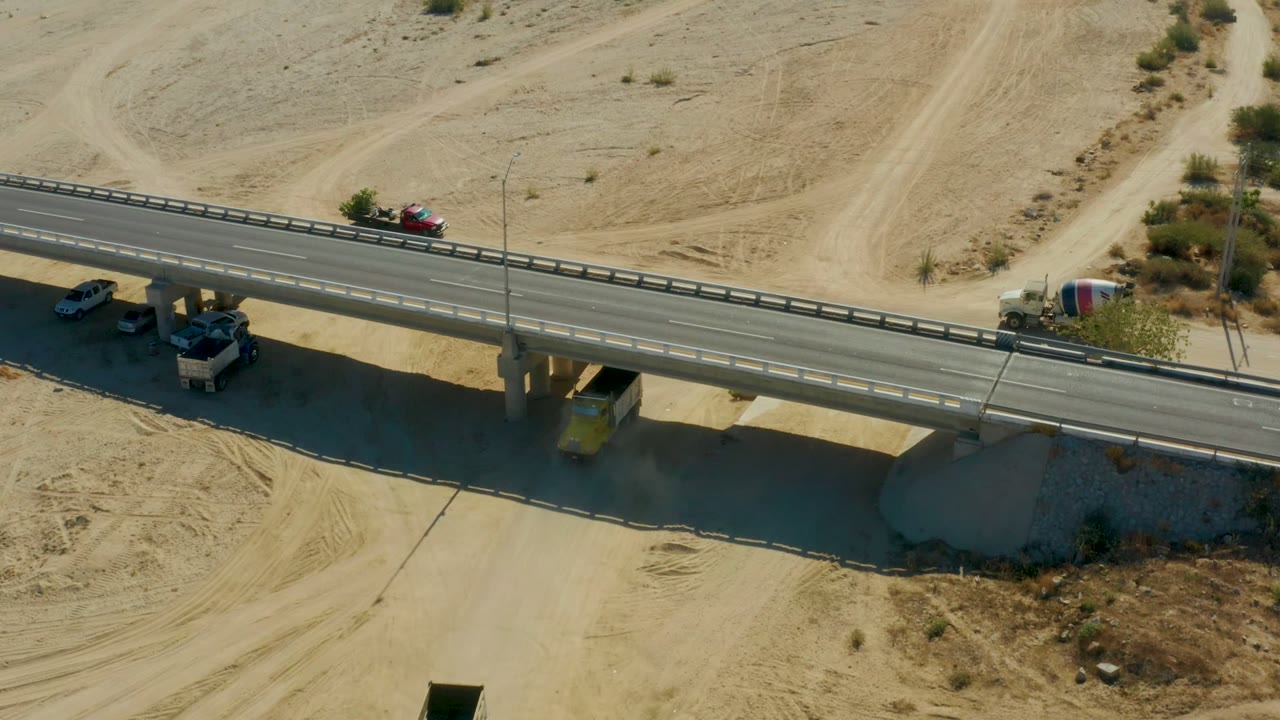 At Chileno Beach Los Cabos, an aerial frame shows a paved bridge over sandy desert with cars, a cement mixer truck, dirt tracks, scrub, and no visible people.