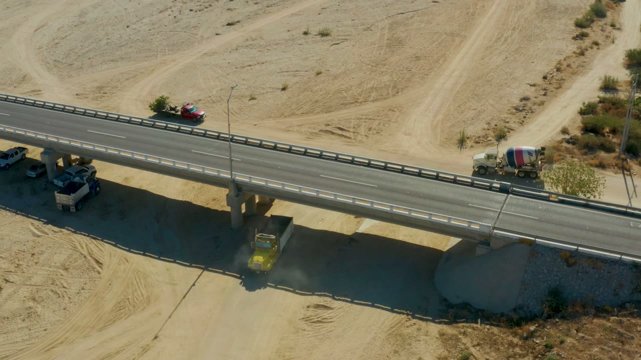 Cement mixer truck and cars move along a bridge over sand.