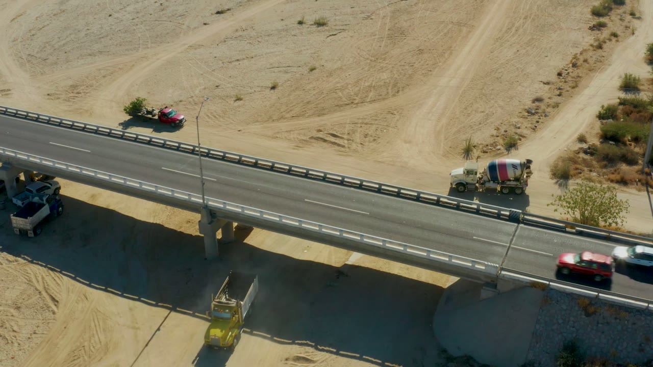 At Chileno Beach Los Cabos, a red car, a white vehicle, and a cement mixer truck are visible on the bridge above pale sand, tire tracks, and scattered desert plants, with no visible people.