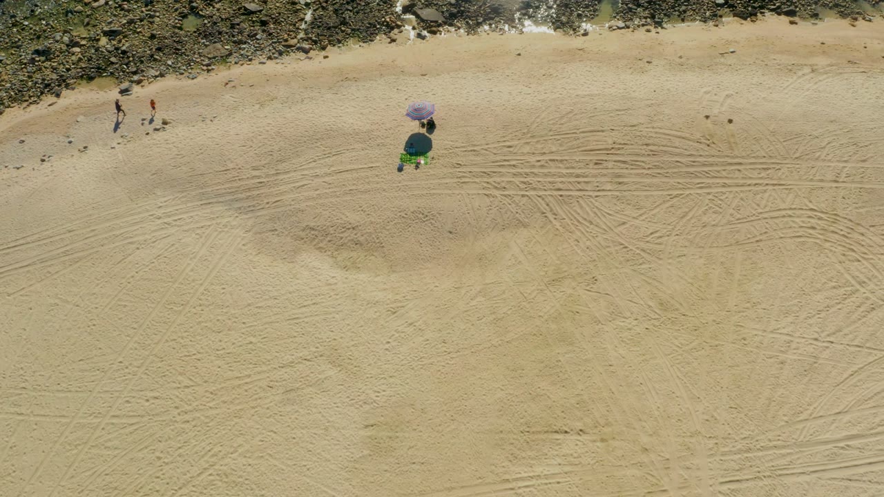 Overhead frame from Chileno Beach Los Cabos showing pale sand, dark rocks along the top edge, two walkers at upper left, and a purple umbrella setup near center.