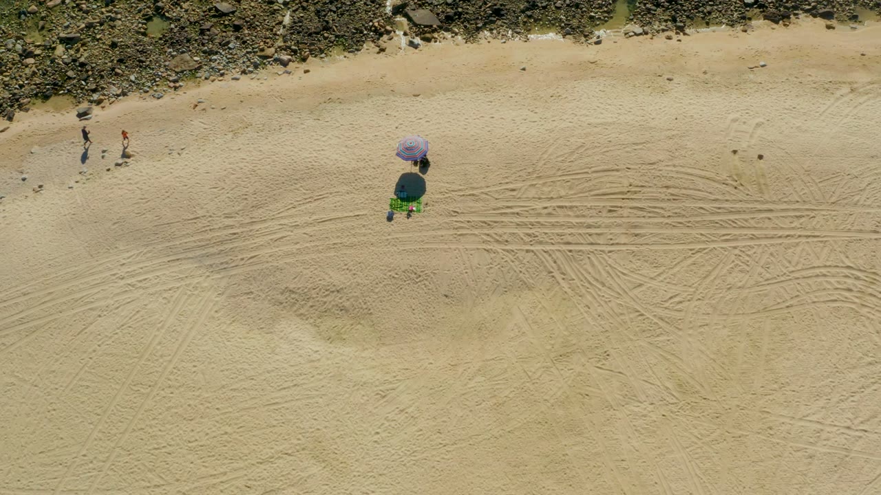Beachgoers walk near tire-marked sand while a purple umbrella sits below rocks.